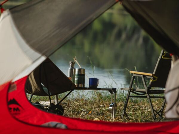 Eine ruhige Aussicht von innen im Zelt mit Blick auf einen Campingplatz am See mit Stuhl und Thermoskanne.