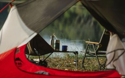 Eine ruhige Aussicht von innen im Zelt mit Blick auf einen Campingplatz am See mit Stuhl und Thermoskanne.